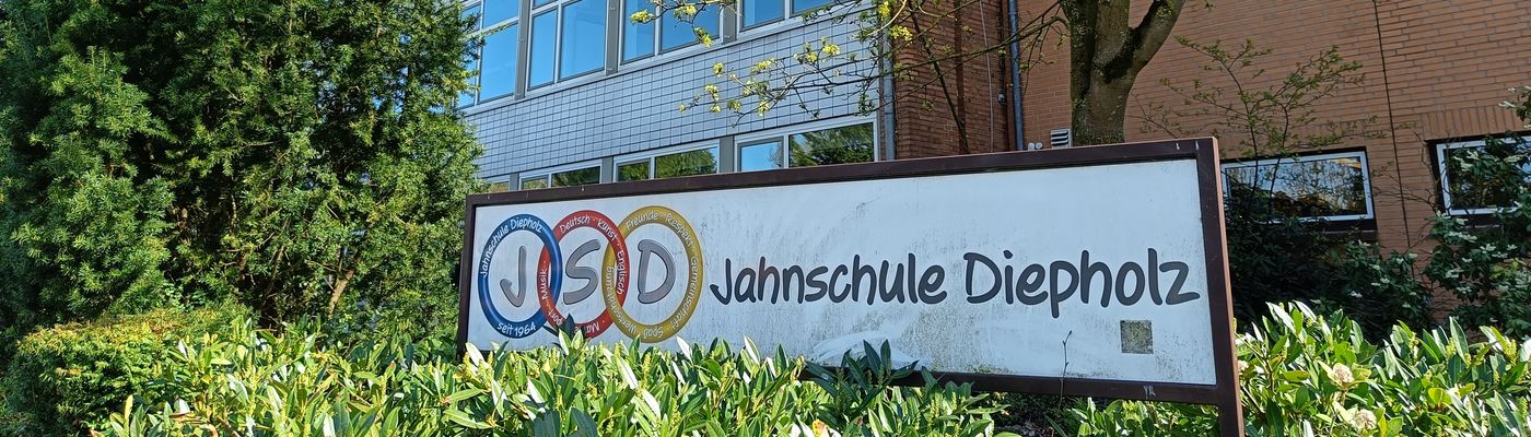 Das Logo der Jahnschule in einem grünen Beet. Das Schulgebäude befindet sich im Hintergrund des Bildes.